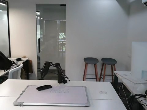 Photo of the real rig: an SO-101 leader arm and follower arm on a desk with a small whiteboard between them, a duster, and a front camera on a stand.